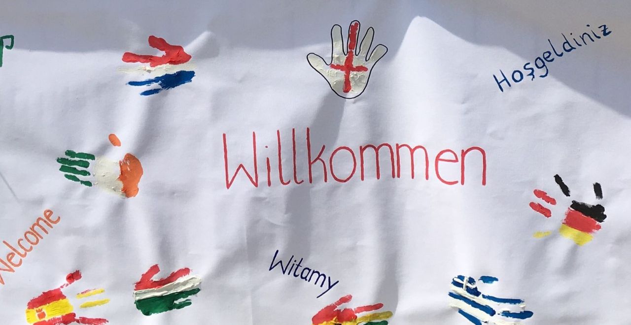 Banner mit bunten Handabdrücken in verschiedenem Flaggendesign. Zusätzlich steht 'Willkommen' auf verschiedenen Sprachen.