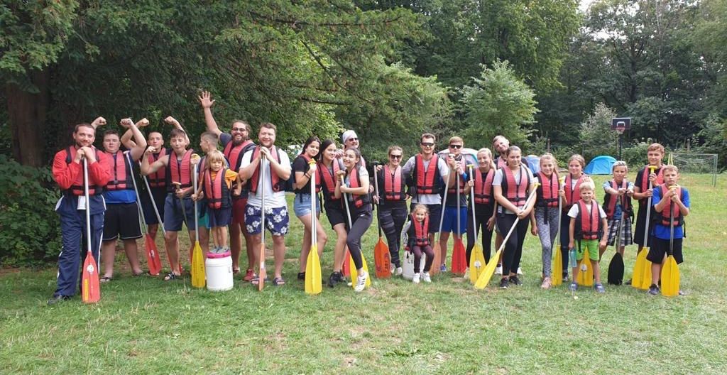 Jugendgruppe steht auf dem Rasen mit Schwimmweste und Paddeln und macht und Gruppenfoto vor dem Kanufahren.