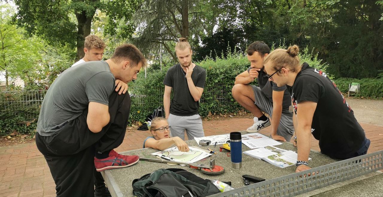 Eine Gruppe von Jugendlichen steht im Park um eine Tischtennisplatte herum und diskutiert über Pläne, die vor ihnen liegen.